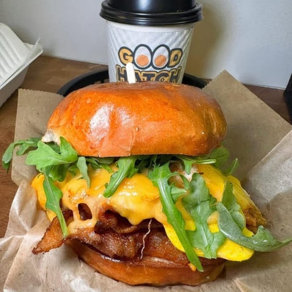 Good Hatch breakfast sandwich with bacon, scrambled eggs, cheese, and arugula, with a branded coffee cup.