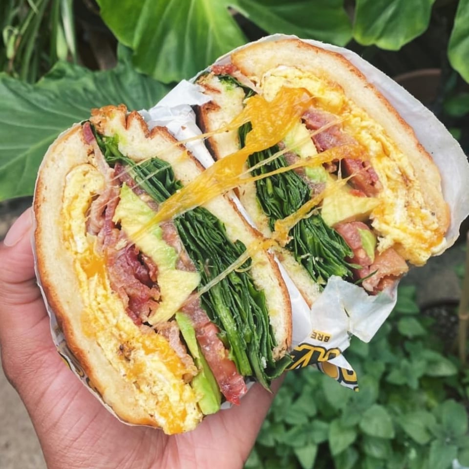 A hand holds a Good Hatch breakfast sandwich, cut in half, revealing egg, bacon, avocado, and greens.