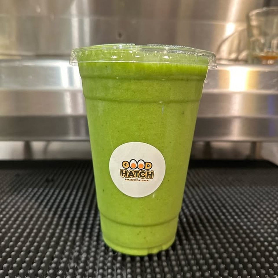 A Good Hatch green smoothie in a clear plastic cup with a lid, on a black textured surface.